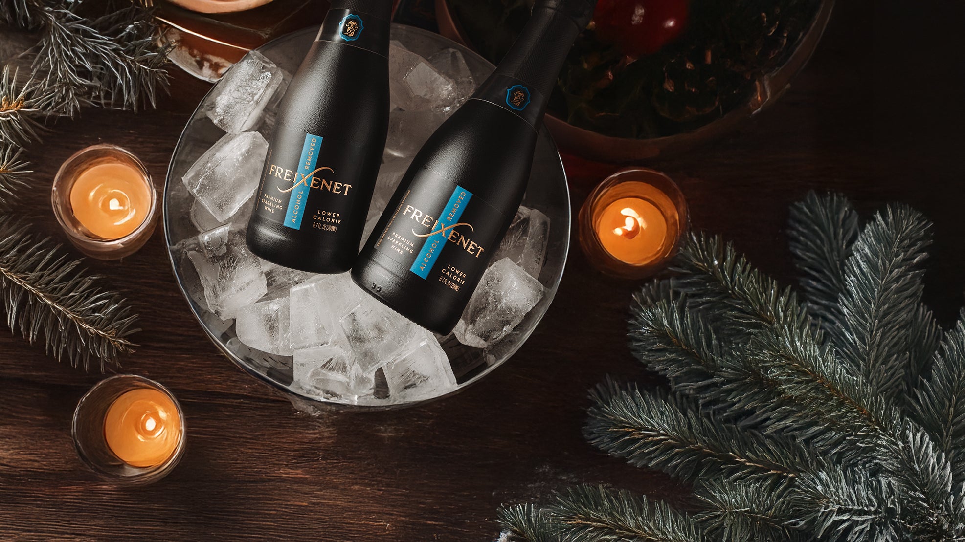Two bottles of Freixenet Alcohol-Removed sparkling wine minis on ice with candles and pine tree decorations on a dark surface.